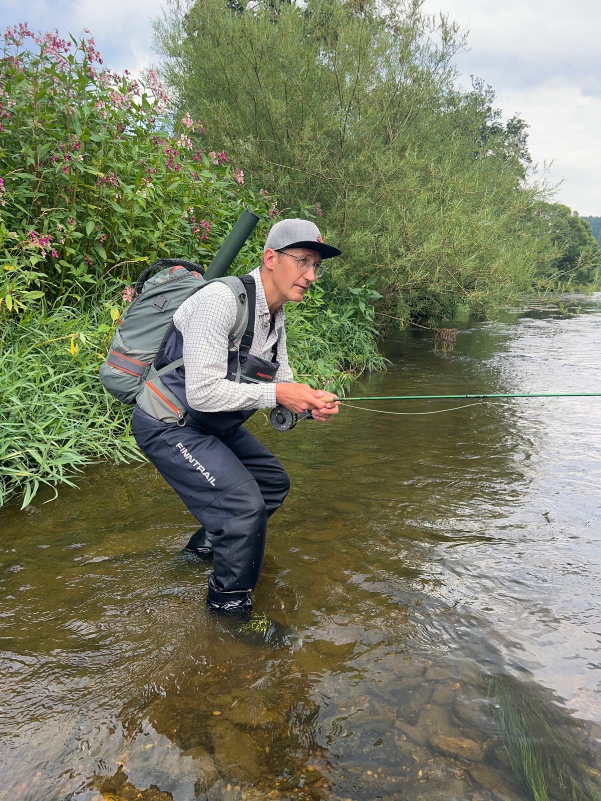 Oleg wading in a river with a fly rod, a backpack and a rod tube on his back